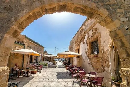 Charming stone houses and outdoor terraces in the historic central square of Marzamemi bathed in sunlight.