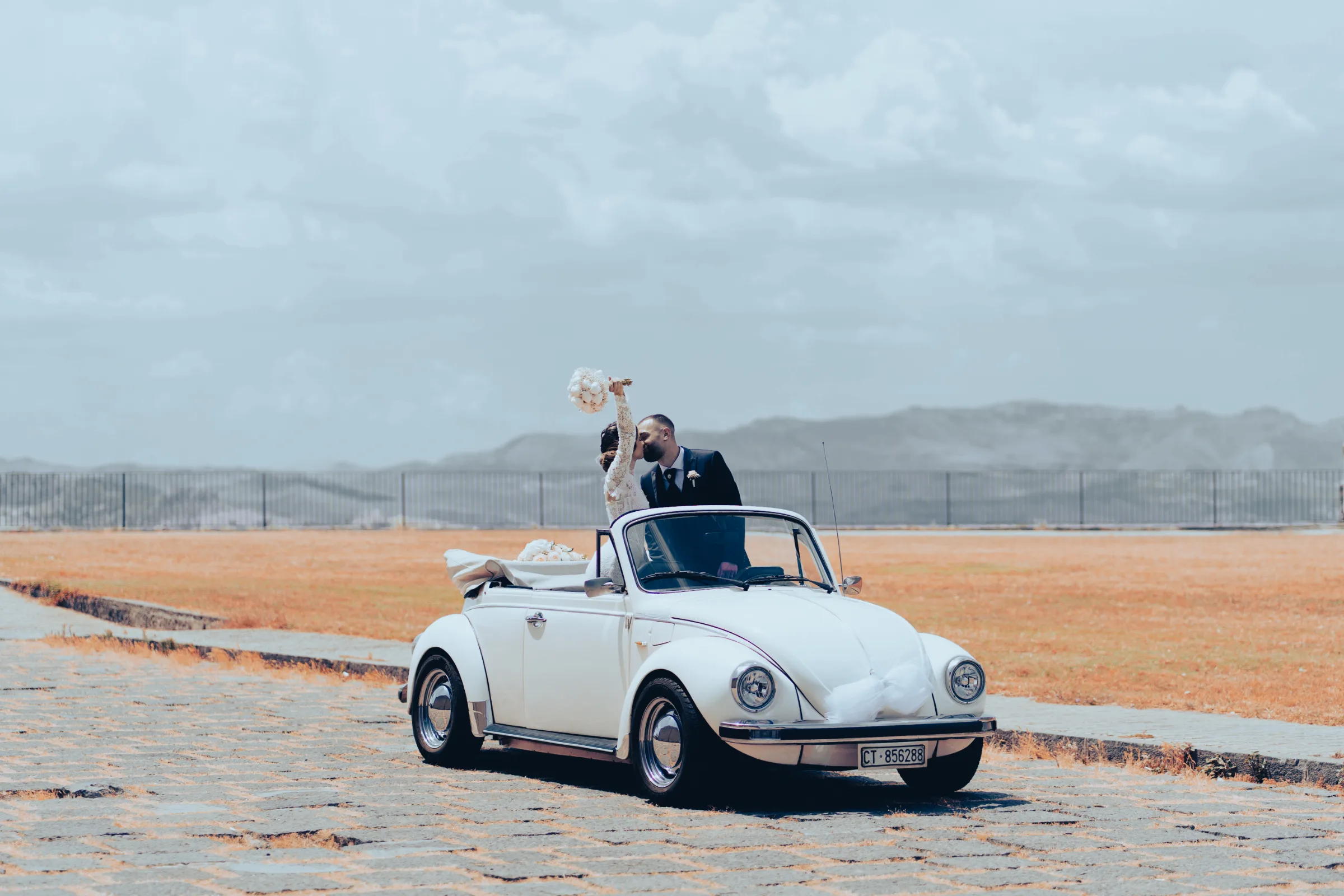 Bride and groom kissing through the open top of a vintage convertible Volkswagen Beetle with scenic Sicilian valley hills in the background